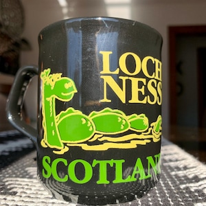 May include: Black ceramic mug featuring a green and yellow illustration of the Loch Ness Monster. The mug has the words "LOCH NESS" and "SCOTLAND" in yellow and green lettering.
