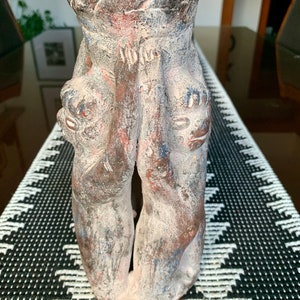 May include: A ceramic vase featuring two figures holding up a rounded top. The figures are painted in shades of brown, blue, and white.