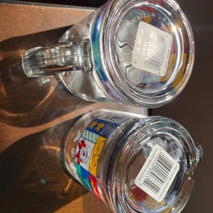 Może przedstawiać: Dwa przezroczyste kubki szklane z kolorowymi wzorami z kresk&oacute;wek i białą naklejką z kodem kreskowym i tekstem "Anchor Hocking Glass Made in USA" na spodzie.