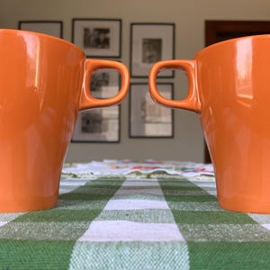 May include: Two orange ceramic mugs with handles, sitting on a green and white checked tablecloth.