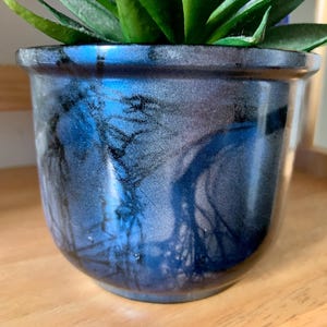 May include: A ceramic plant pot with a glossy, marbled finish in shades of blue, gray, and black. The pot has a rounded shape and a slightly flared rim. Green plant leaves are visible at the top.
