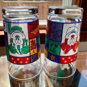 May include: Two clear glass drinking glasses with a winter holiday theme. The glasses feature a patchwork design with Mickey Mouse and Minnie Mouse wearing winter clothing. The glasses have a white snowflake pattern on the bottom.