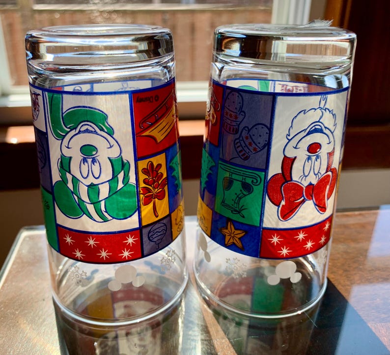 May include: Two clear glass drinking glasses with a winter holiday theme. The glasses feature a patchwork design with Mickey Mouse and Minnie Mouse wearing winter clothing. The glasses have a white snowflake pattern on the bottom.