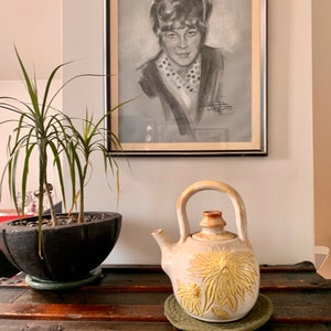 May include: A white ceramic teapot with a floral design and a brown lid. The teapot is sitting on a green woven coaster on a wooden surface. A potted plant and a framed black and white drawing of a woman are in the background.