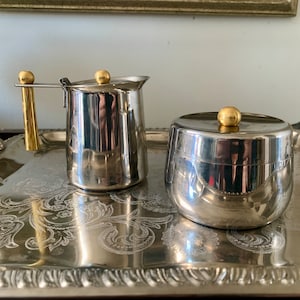 May include: A silver creamer and sugar bowl set with gold accents. The creamer has a handle and a lid. The sugar bowl has a lid. Both pieces are on a silver tray.