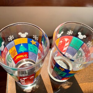 May include: Two clear glass cups with a colorful Christmas design featuring Mickey Mouse and snowflakes. The cups are decorated with a patchwork of red, green, yellow, blue, and white squares.