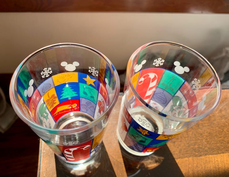 May include: Two clear glass cups with a colorful Christmas design featuring Mickey Mouse and snowflakes. The cups are decorated with a patchwork of red, green, yellow, blue, and white squares.