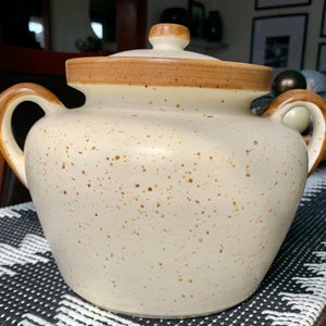 May include: A white ceramic bean pot with a brown lid and speckled brown glaze. The pot has two handles and is perfect for serving a large amount of food.