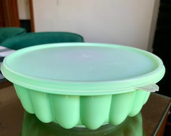 Puede incluir: Un recipiente de almacenamiento redondo de plástico color verde menta claro con un diseño exterior acanalado y festoneado, junto con una tapa circular plana a juego. El recipiente cuenta con una pequeña pestaña transparente en la tapa.