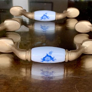 Op de afbeelding: Vijf messing ladehandgrepen met witte keramische centra met blauwe bloemmotieven. De handgrepen hebben een gebogen vorm en zijn gerangschikt op een reflecterend oppervlak, waarbij hun spiegelingen worden getoond. Deze decoratieve hardwarestukken voegen een vleugje elegantie toe.