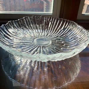 May include: A clear glass serving bowl with a textured, rippled design. The bowl has a wide, shallow shape and a slightly raised center.