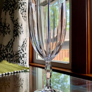 May include: A clear wine glass with a textured, ridged design. The glass stands on a reflective surface, with a window and patterned curtain visible. The glass is designed for serving wine.