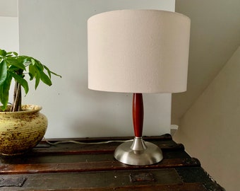 May include: A table lamp with a cream-colored lampshade, a dark wood stem, and a brushed nickel base. The lamp is on a dark wooden trunk. A potted plant with green leaves is to the left of the lamp.