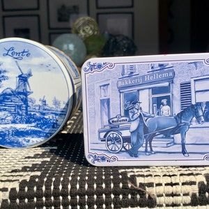 Op de afbeelding: Twee blauwe en witte blikken dozen met Nederlands geïnspireerde ontwerpen. De doos links heeft een windmolen en de tekst "Lente" erop. De doos rechts heeft een bakkerscène met de tekst "Bakkerij Hellema" erop.