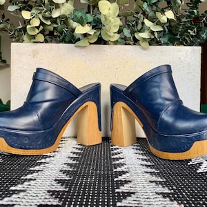 May include: A pair of navy blue slip-on mules featuring a high block heel and a thick platform sole. The upper is constructed from a smooth navy material, while the platform and heel are finished in a light wood grain texture with circular details.