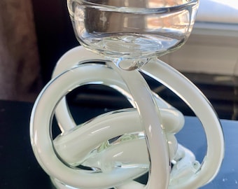 May include: White glass candle holder with a clear glass bowl on top. The holder is shaped like a knot.