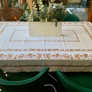 May include: A rectangular white tablecloth with a floral pattern in shades of gold and brown. The tablecloth is set on a wooden table, with a rectangular vase filled with greenery in the center.