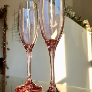 May include: Two pink-tinted champagne flutes. The glasses have a long, thin stem and a broad, circular base. The light catches the glass, creating a shimmering effect. Ideal for a special event or celebration.