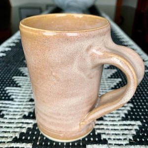 May include: A light brown ceramic mug with a slightly flared rim and a rounded handle. The mug has a speckled glaze with a subtle white and brown pattern.