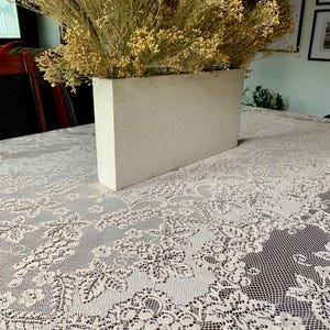 May include: A rectangular, pale concrete planter filled with dried, yellow-toned flowers. The planter sits on a white lace tablecloth with a floral pattern. The scene is set indoors.