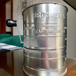 Peut inclure: Un tamis métallique vintage avec une poignée en bois noir. Le tamis est cylindrique avec le texte "BROMWELL'S MEASURING-SIFTER" et des mesures en tasses. Fabriqué aux États-Unis.