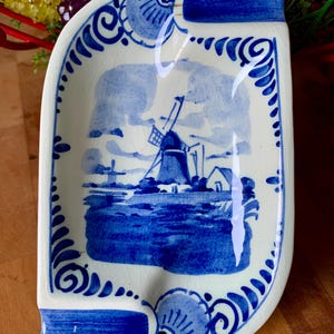 May include: A white and blue ceramic dish with a windmill scene. The dish has a curved shape with a blue border and decorative accents. The windmill scene is painted in shades of blue.