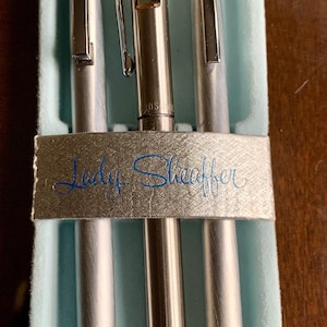 May include: A set of three silver pens in a light blue velvet box. The pens are arranged vertically, with a pen in the centre and two pens on either side. The text "Lady Sheaffer" is printed on the box.