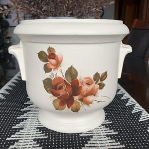 Peut inclure: Pot en céramique blanche avec deux poignées et un motif floral représentant des roses roses et brunes.