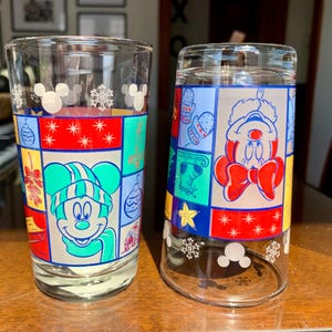 May include: Two clear glass drinking glasses with a Christmas theme. One glass features Mickey Mouse wearing a green hat and scarf, the other features Mickey Mouse wearing a red and white Santa hat. Both glasses have a colorful patchwork design with snowflakes and Mickey Mouse ears.