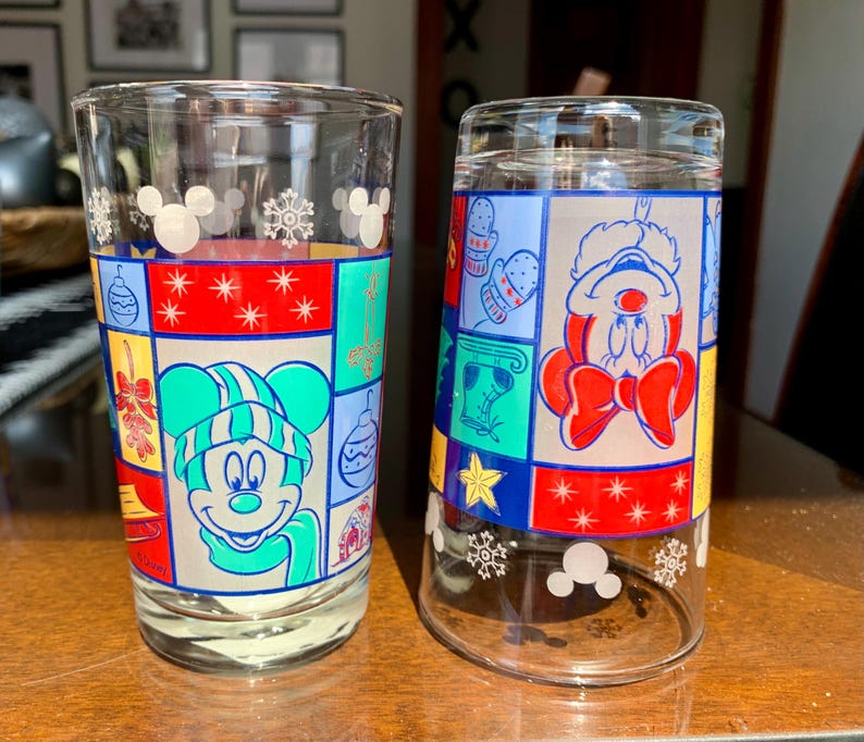 May include: Two clear glass drinking glasses with a Christmas theme. One glass features Mickey Mouse wearing a green hat and scarf, the other features Mickey Mouse wearing a red and white Santa hat. Both glasses have a colorful patchwork design with snowflakes and Mickey Mouse ears.