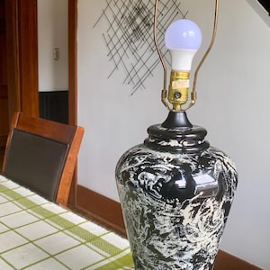 May include: Black and white ceramic table lamp with a bulb. The lamp has a marbled design and a brass-coloured neck. The lamp is on a brown table. Home decor.