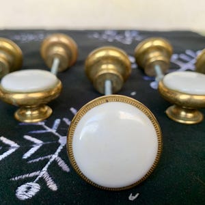 May include: A selection of cabinet knobs with brass and white ceramic elements. The knobs feature a gold-coloured base and a white, round ceramic top. Ideal for furniture and home decor projects.