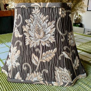 May include: A brown lampshade with a floral pattern in beige and cream. The fabric has a textured, pleated appearance. The lampshade has a flared shape and a scalloped edge.