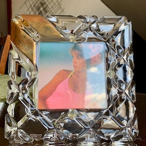 Puede incluir: Un marco de fotos de vidrio transparente y facetado con una abertura cuadrada. El marco tiene un diseño geométrico y un acento dorado en el borde superior. La foto interior muestra a una persona con un top rosa.