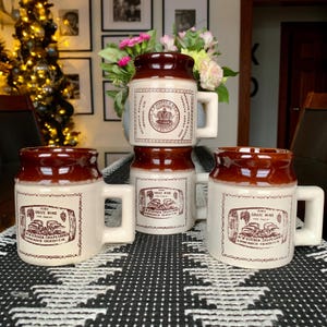 May include: Four brown and white ceramic mugs with a brown rim and a handle. The mugs have a design of a barrel and grapes with the text "Pure Grape Wine For Sale" and "N.W. Kitchen Grape Growers, Wine Maker Grimsby, Ont."