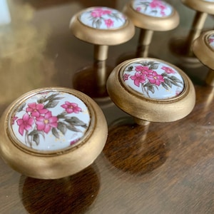 May include: A collection of gold-toned cabinet knobs. Each knob features a white ceramic face with a pink floral design and a gold-colored rim. The knobs are set against a blurred wooden surface.
