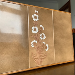May include: A brown wooden box with three panels, each featuring a white floral design. The box has a glossy finish and a simple, elegant design.