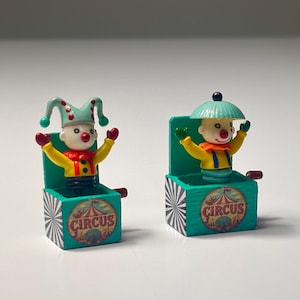 Op de afbeelding: Twee miniatuur speeldoosjes met clowns. De linker clown draagt een mintgroene narrenmuts met rode stippen. De rechter clown draagt een mintgroene hoed. Beide doosjes zijn turquoise met zwart-witte strepen en een rond circuslogo op de voorzijde.