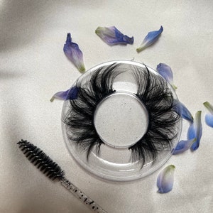 Puede incluir: Un par de pestañas postizas negras en una bandeja de plástico transparente con pétalos de flores azules esparcidos a su alrededor. También se incluye un aplicador de pestañas de plástico transparente con un cepillo negro.