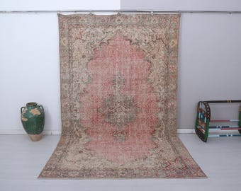May include: A large, rectangular, vintage rug with a central medallion design in shades of pink, beige, and brown. The rug is displayed against a white wall, with a green vase and a decorative bench in the background.