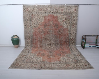 Puede incluir: Una alfombra vintage grande y rectangular con un diseño floral ornamentado. La alfombra presenta un medallón central en tonos rojos y marrones, rodeado por un borde beige con patrones intrincados. La alfombra se exhibe sobre un suelo claro.
