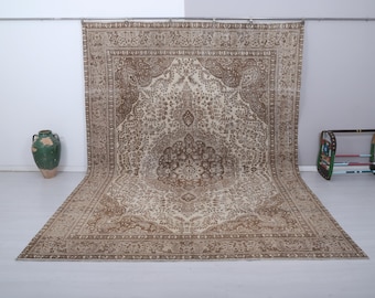 May include: A large, rectangular, vintage rug with an intricate floral pattern in shades of beige and brown. The rug is displayed on a white floor, with a green vase and a small wooden shelf in the background.