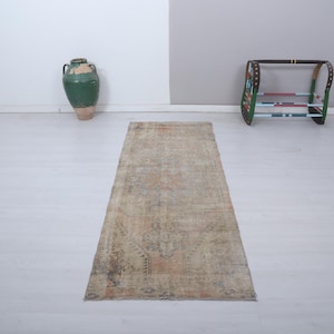 May include: A rectangular, distressed beige rug with a faded, patterned design. The rug is placed on a light-coloured floor, with a green vase and a decorative rack in the background. The rug's design features muted tones of brown and orange.