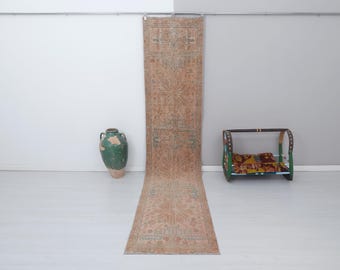 May include: A long, rectangular rug with a faded, floral pattern in shades of pink, beige, and blue. The rug is hanging on a wall, with a green ceramic vase and a wooden bench in the foreground.