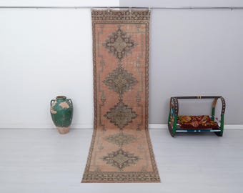 May include: A long, narrow rug with a faded pink background and intricate dark green geometric patterns. The rug is hanging on a wall with a white background.