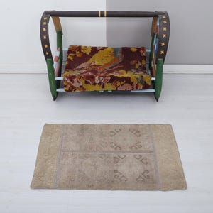 Può includere: Un'altalena decorativa in legno con seduta fantasia e tappeto abbinato. L'altalena ha una barra superiore marrone, supporti laterali verdi e marroni e una seduta fantasia con tonalità rosse, gialle e marroni. Il tappeto è rettangolare e beige.
