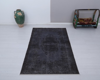 Puede incluir: Una alfombra rectangular gris oscuro con un patrón desgastado. La alfombra está colocada sobre un suelo blanco, con un jarrón verde y un pequeño columpio decorativo en el fondo. El diseño de la alfombra presenta áreas descoloridas.
