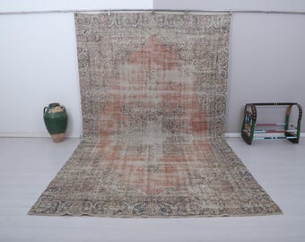 Puede incluir: Una alfombra grande, rectangular y desgastada con un diseño central naranja descolorido y un borde ornamentado. La alfombra se exhibe contra una pared blanca, con un jarrón verde y una estantería decorativa cerca. Las dimensiones de la alfombra no son visibles.