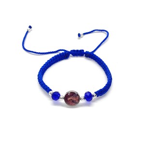 May include: A blue braided bracelet with a central circular charm featuring a religious image. The bracelet includes two blue faceted beads and silver-colored metal accents. The adjustable cord is also blue.