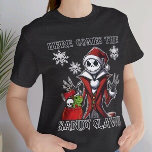 Pu&ograve; includere: Una maglietta nera con una grafica bianca di Jack Skellington vestito da Babbo Natale. La grafica dice "Here Comes The Sandy Claws".